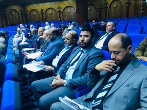 Group of professionals seated in blue auditorium chairs, some reading documents during a conference event in a formal setting.