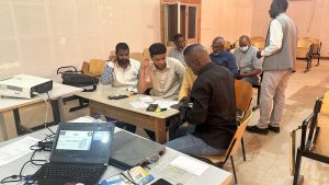 Group of men seated at a table in a classroom, examining papers as a man in a white shirt points to something at the center.