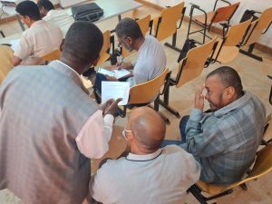 Group of men in a classroom collaborating over papers; one man writes on a pad while others review documents.