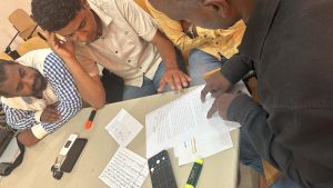 Four men sit around a table reviewing handwritten notes and a printed document during a collaborative discussion; a hand points to the paper while others look on.