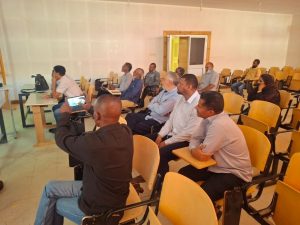 Group of adults seated in a classroom-like lecture hall, attentively watching a presenter off-camera; a man in the foreground records with a smartphone.
