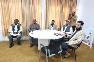 Six men sit around a round white table in a sunlit room with gold curtains; a photographer stands at the back taking pictures