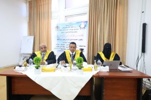 Panel of three graduates in black gowns with yellow sashes seated at a conference table, a banner behind them and a laptop on the right; middle speaker gesturing as he talks.