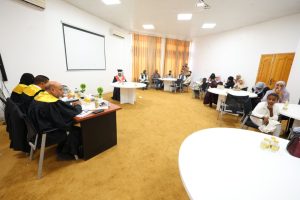 Formal ceremony in a bright room: graduates in black gowns with yellow sashes sit at a table on the left while officials at a round white table address them; attendees sit at tables along the right wall under orange curtains.