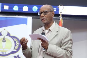 Older man in a light suit speaks into a handheld microphone, holding notes in front of a banner with a crest logo behind him.