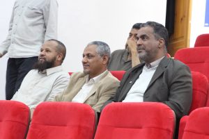 Three men in casual business attire seated in red auditorium chairs, attentively watching a presentation.