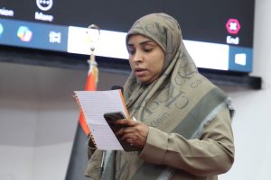 Woman in a beige hijab and coat reads from a lined notebook while holding a phone and speaking into a microphone at an event backdrop.