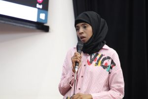 Woman in a black hijab speaking into a handheld microphone on stage, wearing a pink striped shirt with colorful graphic.