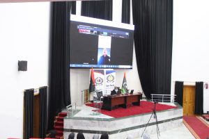 Speaker at a desk on a raised stage with a large screen showing a video call above, flanked by flags and banners in a formal auditorium.
