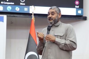 Man with gray beard speaks into a handheld microphone, standing beside a red-and-black flag during a presentation.