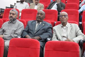 Three men seated in red theater seats, attentively watching an event in a formal setting.