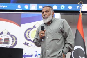 Bearded man in a gray shirt holding a microphone on stage, with banners and a Libyan flag behind him.