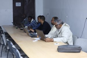 Six men sit along a long conference table, each working on their laptops in a meeting room.