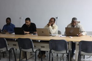 Four men sit at a long conference table with laptops, focused on their screens in a meeting room.