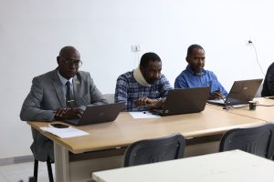 Three men sit at a conference table with laptops; a suited man on the left, a man with a neck brace in the center, and a man in a blue shirt on the right.