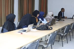 Group of women wearing hijabs sit along a long table with laptops, a man works at the far end in a classroom setting