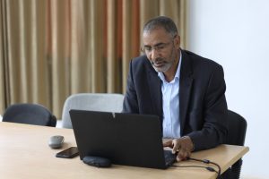 Middle-aged man in a pinstriped suit typing on a laptop at a wooden table in a meeting room with beige curtains behind him.
