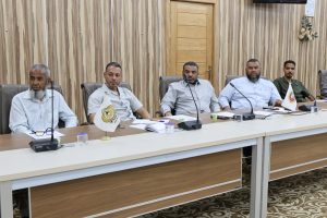 Five men sit at a long conference table in a wood-paneled meeting room, with microphones, papers, and small flags in front of them.