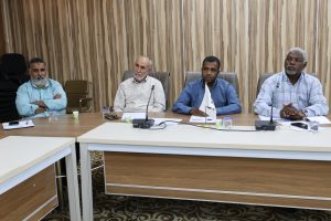 Four men sit at a conference table in a meeting room with microphones and papers before them.