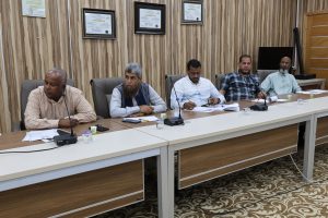 Five men sit at a long conference table in a meeting room with wood-paneled walls and framed certificates, microphones in front of them.