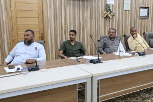 Four men sit at a long conference table in a wood-paneled room, each with papers and a microphone in front of them, suggesting a formal meeting or panel discussion.