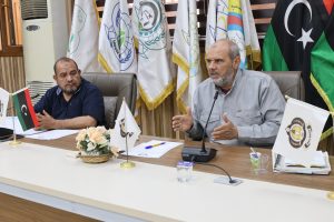 Two men sit at a conference table with flags behind them; one speaks into a microphone while the other listens.