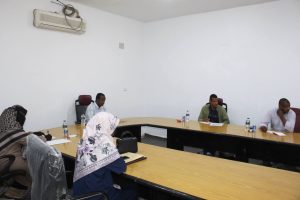 People seated around a large U-shaped conference table in a bright meeting room, with water bottles, papers, and notebooks; bare white walls and an air conditioner mounted high on the wall.