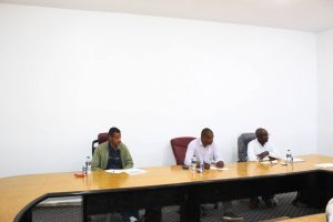 Four men sit at a long wooden conference table in a bright white room, with papers and water bottles in front of them.