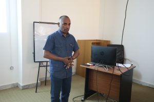 Man in a denim shirt stands at the front of a classroom, speaking beside a desk with a laptop and tangled cables.