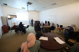 Instructor presenting to a seated audience in a meeting room with a projector screen at the front.