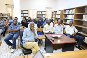 Group of adults seated in a library classroom listening to a presentation next to bookshelves and a projector on a table front.