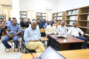 Group of adults seated in a library classroom, facing forward as a projector sits on a table at the front. Bookshelves line the back wall.