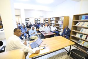 Adults seated in a library classroom listening to a presenter at a laptop-enabled table in front of the room.