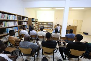 Group of men seated in a library classroom listening to a presenter at the front, with a projector screen glowing in the background.