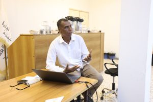 Man in a white shirt seated at a wooden table with an open laptop, gesturing as he speaks.