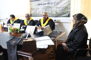 Panel of academics in black robes with yellow sashes seated at a conference table; a woman in a hijab sits on the right, with laptops and drinks visible, and a banner with Arabic text behind them.