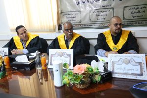 Three men in black academic gowns with yellow stoles sit at a conference table during a formal ceremony, with an Arabic banner behind them.