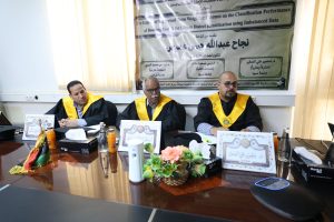 Three men in black academic robes with yellow sashes sit at a conference table, with a large Arabic banner behind them and bottles, flowers, and boxes on the table.