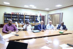 Group of men in a conference room seated around a long wooden table, with awards shelves and flags behind them.
