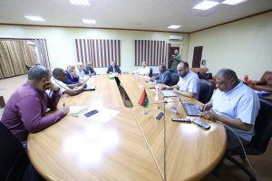 Group of men in a boardroom seated around a large wooden table, flags in the center, taking notes.