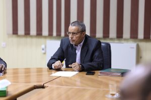 Man in a dark suit and glasses sits at a conference table, holding a yellow pencil during a meeting with papers and a phone nearby.