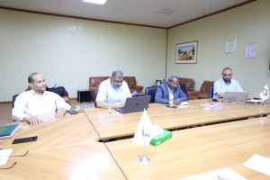 Four men sit around a large wooden conference table with laptops and documents in a light-paneled room.