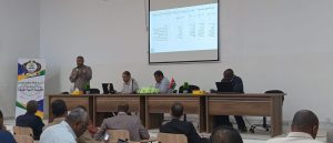 Panel discussion in a conference room with five men at a long table and a projector screen displaying Arabic slides above them
