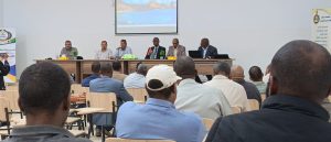 Panel of six men seated at a long table at the front of a conference room addressing an audience; projector screen shows a landscape image.