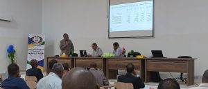 Speaker stands at a panel desk giving a lecture while audience watches in a conference room with a projected slide on screen.