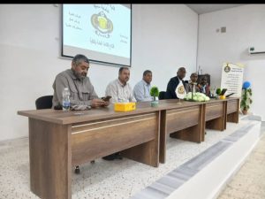 Six men sit at a long wooden panel table on a stage, microphones in front, with a projected Arabic slide behind them.