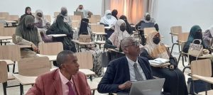 Classroom with attendees wearing hijabs and masks; two men in the front use a laptop.