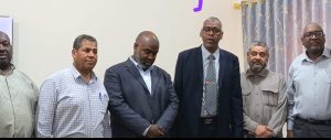 Six men in business attire pose for a group photo in a conference room, standing side by side indoors.