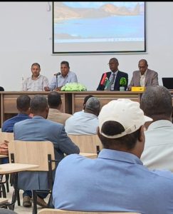 Panel of four men seated at a long table on a stage in a lecture hall, addressing an audience; a large screen with a landscape image is projected behind them.