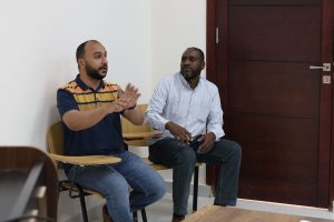 Two men seated and talking in a room; the left man gestures with his hands while the right man listens, against a white wall and dark wooden door.